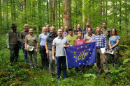 Eine Gruppe Menschen, die eine blaue Flagge halten, steht im Wald und grinst in die Kamera