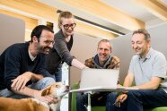 Vier Mitarbeitende sitzen um einen Tisch und schauen zusammen in einen Laptop. Ein Hund ist bei ihnen