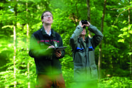 Zwei Männer in Uniform der Forstverwaltung stehen im Wald, der eine schreibt etwas auf, während der andere mit einem Fernglas in die Kronen blickt