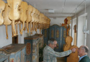 Das Foto zeigt einen Raum in dem zahlreiche Geigen aus hellem Holz mit dem Hals nach unten an der Decke aufgehängt sind. Ansonsten befinden sich bemalte Bauernschränke und zwei Herren in dem Raum. 