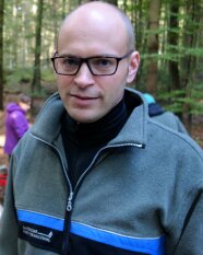 Junger Mann mit Forstuniformjacke, Glatze und dunkler Brille steht in einem Wald.