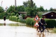 Überflutete Straße: Ein Fahrradfahrer versucht durch das Wasser zu fahren. Im Hintergrund steht ein Mann knietief im Wasser.