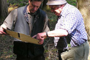 Älterer Mann mit Hut und mittelalter Mann mit Unterlagen in der Hand beraten sich im Wald.