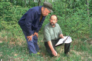 Das Bild zeigt zwei Männer, die auf einer Lichtung im Wald stehen. Einer der Männer ist blau gekleidet und steht in einer gebeugten Haltung, während er dem anderen knieenden Mann zuhört.