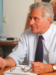 Das Bild zeigt einen Mann mit grauen Haaren, der  ein blaues Hemd und eine dunkelblaue Krawatte an hat. Der Mann sitzt an einem Schreibtisch auf dem eine Brille und Papiere liegen.