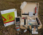 Das Bild zeigt einen offenen Holzkoffer und ein Buch mit grünem Titelbild auf einem braunen Untergrund.