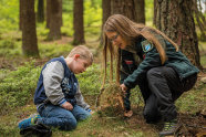 Eine junge Frau mit Brille und Amtsuniform kniet auf dem Boden und zeigt einem kleinen Jungen, was sich unter der Moosschicht des Waldes verbirgt