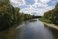 Das Foto zeigt einen Fluss, der links mit Laubbäumen gesäumt ist und rechts befindet sich ein kleiner Streifen Steinstrand entlang des Flusses.