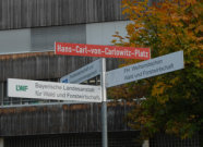 Das Bild zeigt ein rotes Strassenschild mit der Aufschrift "Hans-Carl-von-Carlowitz-Platz". Unter dem Schild sind drei Wegweiser angebracht, die in drei verschiedene Richtungen zeigen.