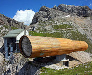 Das Bild zeigt ein überdimensionales Fernrohr aus Holz, das an einer Felskante im Hochgebirge befestigt ist. Im Hintergrund sieht man einen Berg und eine Bergstation.