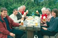 Das Foto zeigt 8 Personen, die an einem Holztisch auf zwei gegenüberliegenden Bänken Brotzeit machen. Sie heben ihre Getränke hoch um in die Kamera zu "Prosten".
