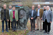 Sieben ältere Männer stehen um ein Denkmal in Form eines Steines und posieren für das Bild