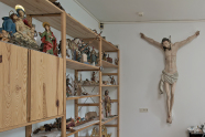Ein weiß gestrichener Raum mit einem Jesuskreuz und links davon Regale, in denen Holzfiguren stehen