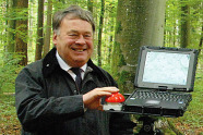 Minister Helmut Brunner im Anzug steht im Wald neben einem Computer und drückt auf einen roten Buzzer.