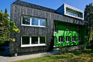 Rückenacnsicht eines grauen Hauses mit grün eingerahmter Fensterfront