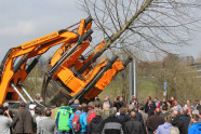 Ein Baum wird mit einer großen Maschine gepflanzt. Menschen stehe davor und schauen zu.