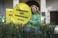 Frau mit grüner Jacke hält gelb-grünes rundes Transparent mit der Schrift "Buchenwälder statt Industrieforste" hoch.