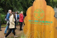 Eine Gruppe Menschen in Regenjacken spaziert an einem Schild aus Holz mit der Aufschrift Kur- und Heilwald vorbei