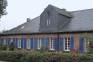 Ein lang gestrecktes, geducktes Haus aus Backsteinen mit Schieferplatten auf dem Dach und einem gewaltigen Hirschhaupt an der Dachgaube und blauen Fensterläden